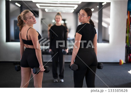 Workout session featuring two women with dumbbells and a trainer in a gym setting, emphasizing strength and fitness 133385626
