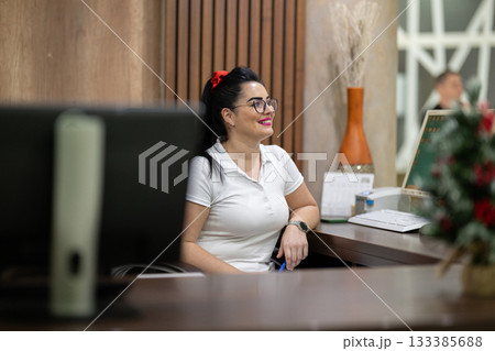 Female receptionist wearing glasses and white uniform smiling at the front desk of a spa and wellness center, representing hospitality, customer care, and professional service. Female receptionist wearing glasses and white uniform smiling at the front desk of a spa and wellness center, representing hospitality, customer care, and professional service. 133385688