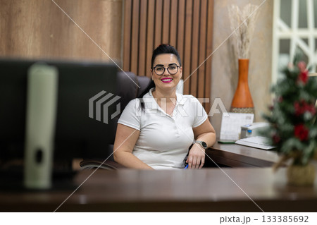 Female receptionist wearing glasses and white uniform smiling at the front desk of a spa and wellness center, representing hospitality, customer care, and professional service. 133385692