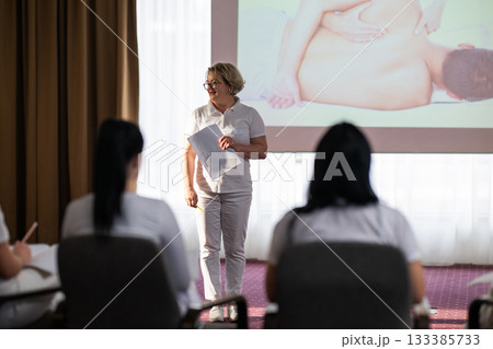 Group of students attending a professional massage therapy training class with instructor presenting on a projector screen, focused on anatomy and therapeutic techniques. 133385733