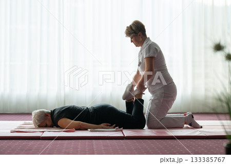 Professional female physiotherapist helping an older woman stretch her legs while lying face down on a mat. Rehabilitation and physiotherapy concept focusing on mobility recovery, flexibility, and Professional female physiotherapist helping an older woman stretch her legs while lying face down on a mat. Rehabilitation and physiotherapy concept focusing on mobility recovery, flexibility, and 133385767