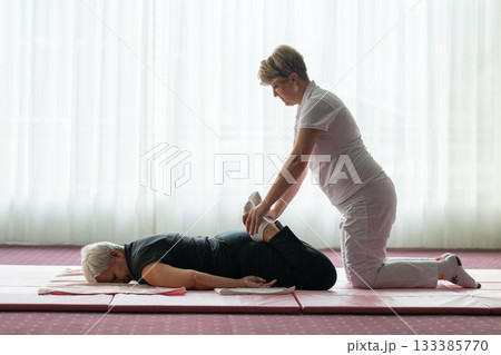 Professional female physiotherapist helping an older woman stretch her legs while lying face down on a mat. Rehabilitation and physiotherapy concept focusing on mobility recovery, flexibility, and Professional female physiotherapist helping an older woman stretch her legs while lying face down on a mat. Rehabilitation and physiotherapy concept focusing on mobility recovery, flexibility, and 133385770