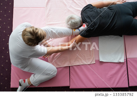A physiotherapist assisting an older woman with a shoulder and arm stretch while lying on a mat. Rehabilitation and physiotherapy session concept focusing on mobility, flexibility, pain relief, and 133385792