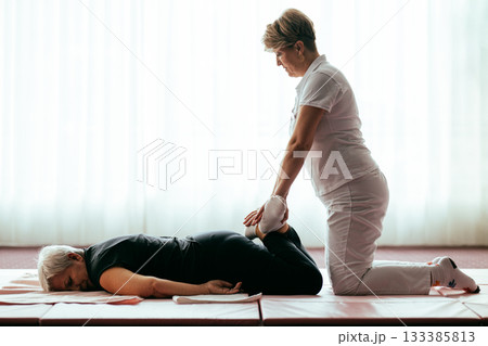 Professional female physiotherapist helping an older woman stretch her legs while lying face down on a mat. Rehabilitation and physiotherapy concept focusing on mobility recovery, flexibility, and 133385813