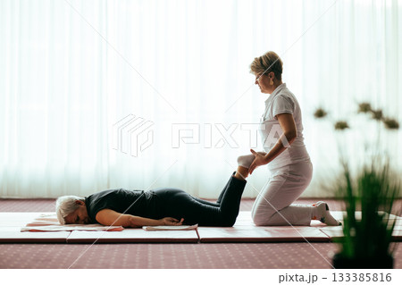 Professional female physiotherapist helping an older woman stretch her legs while lying face down on a mat. Rehabilitation and physiotherapy concept focusing on mobility recovery, flexibility, and Professional female physiotherapist helping an older woman stretch her legs while lying face down on a mat. Rehabilitation and physiotherapy concept focusing on mobility recovery, flexibility, and 133385816