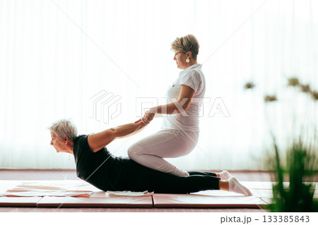 Professional female physiotherapist supporting an older woman during a therapeutic stretching exercise on a mat. Rehabilitation and physiotherapy concept focused on mobility, flexibility, recovery 133385843