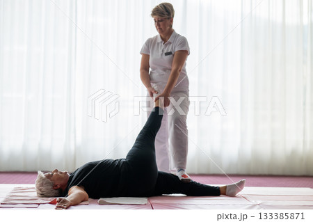 Professional female physiotherapist assisting an older woman with a leg stretch while lying on a mat during a rehabilitation session. Concept of physiotherapy, mobility recovery, flexibility, and 133385871