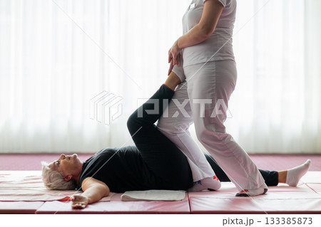Professional female physiotherapist assisting an older woman with a leg stretch while lying on a mat during a rehabilitation session. Concept of physiotherapy, mobility recovery, flexibility, and 133385873