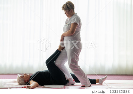 Professional female physiotherapist assisting an older woman with a leg stretch while lying on a mat during a rehabilitation session. Concept of physiotherapy, mobility recovery, flexibility, and Professional female physiotherapist assisting an older woman with a leg stretch while lying on a mat during a rehabilitation session. Concept of physiotherapy, mobility recovery, flexibility, and 133385888