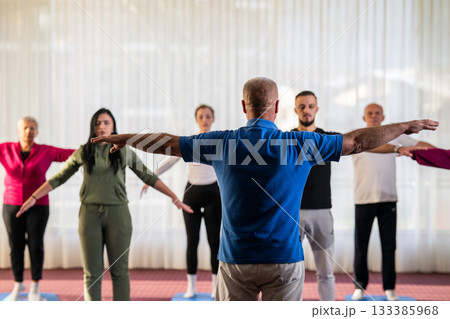 Instructor leading a diverse group of people during a fitness training session with stretching exercises on mats in a bright wellness center 133385968