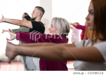 Group of people of different ages participating in a guided fitness or rehabilitation session indoors, standing on yoga mats with hands behind heads. Group of people of different ages participating in a guided fitness or rehabilitation session indoors, standing on yoga mats with hands behind heads. 133386118
