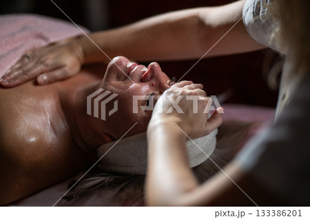 Close up of a mature woman receiving a relaxing facial massage from a professional female therapist in a modern spa. The client lies on a treatment bed with a towel and headband, enjoying skincare Close up of a mature woman receiving a relaxing facial massage from a professional female therapist in a modern spa. The client lies on a treatment bed with a towel and headband, enjoying skincare 133386201