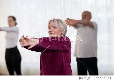 Active elderly woman in sportswear stretching her arms forward during a group fitness or rehabilitation session. Concept of healthy aging, wellness, flexibility, and active senior lifestyle. Active elderly woman in sportswear stretching her arms forward during a group fitness or rehabilitation session. Concept of healthy aging, wellness, flexibility, and active senior lifestyle. 133386224