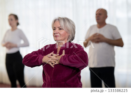 Active elderly woman in sportswear stretching her arms forward during a group fitness or rehabilitation session. Concept of healthy aging, wellness, flexibility, and active senior lifestyle. Active elderly woman in sportswear stretching her arms forward during a group fitness or rehabilitation session. Concept of healthy aging, wellness, flexibility, and active senior lifestyle. 133386225