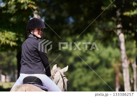 Smiling woman riding a horse in nature, equestrian sport, horseback riding in the countryside, outdoor activity, healthy lifestyle, enjoying nature, sunny day. 133386257