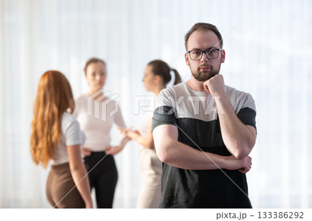 Portrait of a confident young man wearing glasses and smiling while standing with his colleagues in the background during an informal business meeting. Portrait of a confident young man wearing glasses and smiling while standing with his colleagues in the background during an informal business meeting. 133386292
