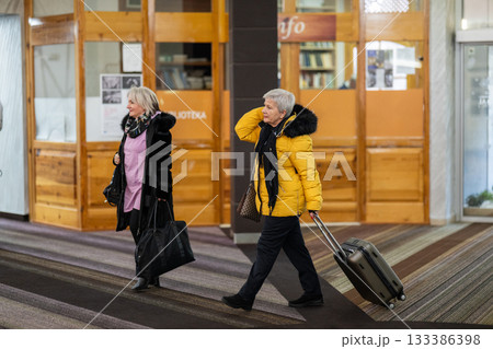 Two older women dressed in winter clothing standing in a hotel lobby and having a friendly conversation next to their luggage. Concept of travel, friendship, and leisure lifestyle for seniors. 133386398