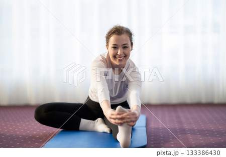Focused woman stretching her leg while sitting on a yoga mat in a bright indoor space. Concept of wellness, flexibility, mindfulness, and healthy lifestyle through exercise and relaxation. Focused woman stretching her leg while sitting on a yoga mat in a bright indoor space. Concept of wellness, flexibility, mindfulness, and healthy lifestyle through exercise and relaxation. 133386430