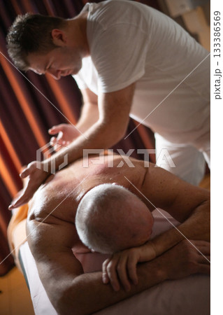 Professional therapist applying deep tissue massage techniques on a male client lying on massage table with towel, therapeutic bodywork for muscle pain relief and relaxation. 133386569