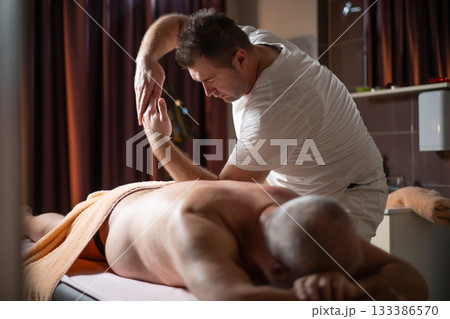 Professional therapist applying deep tissue massage techniques on a male client lying on massage table with towel, therapeutic bodywork for muscle pain relief and relaxation. 133386570