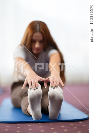 Focused woman stretching her leg while sitting on a yoga mat in a bright indoor space. Concept of wellness, flexibility, mindfulness, and healthy lifestyle through exercise and relaxation. 133386576