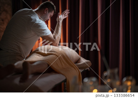 Professional male massage therapist performing deep tissue back massage on an older man lying on a massage table covered with a towel. Relaxing wellness treatment in a modern spa room with warm light Professional male massage therapist performing deep tissue back massage on an older man lying on a massage table covered with a towel. Relaxing wellness treatment in a modern spa room with warm light 133386654