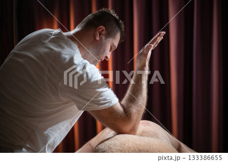 Professional male massage therapist performing deep tissue back massage on an older man lying on a massage table covered with a towel. Relaxing wellness treatment in a modern spa room with warm light Professional male massage therapist performing deep tissue back massage on an older man lying on a massage table covered with a towel. Relaxing wellness treatment in a modern spa room with warm light 133386655
