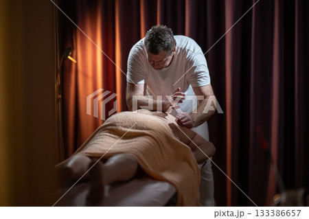 Professional male massage therapist performing deep tissue back massage on an older man lying on a massage table covered with a towel. Relaxing wellness treatment in a modern spa room with warm light Professional male massage therapist performing deep tissue back massage on an older man lying on a massage table covered with a towel. Relaxing wellness treatment in a modern spa room with warm light 133386657