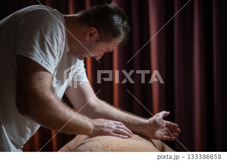 Professional male massage therapist performing deep tissue back massage on an older man lying on a massage table covered with a towel. Relaxing wellness treatment in a modern spa room with warm light Professional male massage therapist performing deep tissue back massage on an older man lying on a massage table covered with a towel. Relaxing wellness treatment in a modern spa room with warm light 133386658