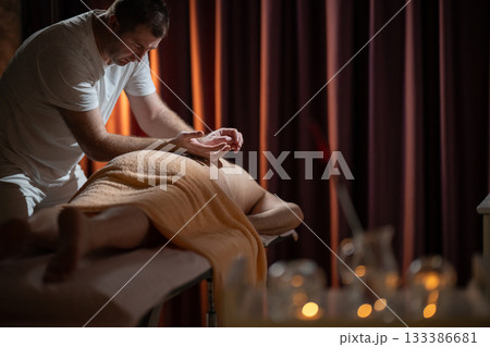 Professional male massage therapist performing deep tissue back massage on an older man lying on a massage table covered with a towel. Relaxing wellness treatment in a modern spa room with warm light Professional male massage therapist performing deep tissue back massage on an older man lying on a massage table covered with a towel. Relaxing wellness treatment in a modern spa room with warm light 133386681