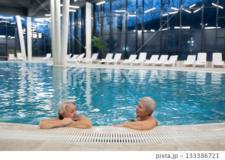 Two smiling senior women enjoying leisure time in an indoor swimming pool, leaning on the poolside. Concept of friendship, wellness, active lifestyle, relaxation, and healthy aging. 133386721