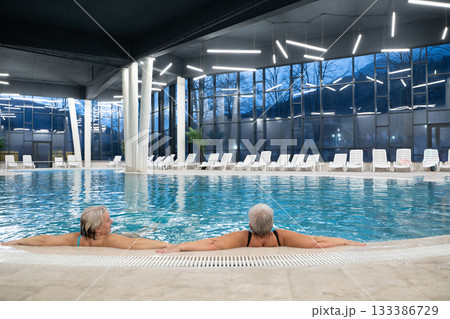 Two senior women enjoying relaxation while leaning on the edge of a modern indoor swimming pool in the evening. Concept of wellness, healthy lifestyle, spa, leisure, and friendship for older adults. Two senior women enjoying relaxation while leaning on the edge of a modern indoor swimming pool in the evening. Concept of wellness, healthy lifestyle, spa, leisure, and friendship for older adults. 133386729