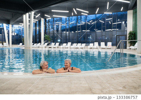 Two smiling senior women enjoying leisure time in an indoor swimming pool, leaning on the poolside. Concept of friendship, wellness, active lifestyle, relaxation, and healthy aging. Two smiling senior women enjoying leisure time in an indoor swimming pool, leaning on the poolside. Concept of friendship, wellness, active lifestyle, relaxation, and healthy aging. 133386757
