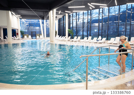 Elderly woman sitting by the edge of a modern indoor spa pool, enjoying the relaxing water jets. Wellness and relaxation concept in a luxurious spa or wellness center. Elderly woman sitting by the edge of a modern indoor spa pool, enjoying the relaxing water jets. Wellness and relaxation concept in a luxurious spa or wellness center. 133386760