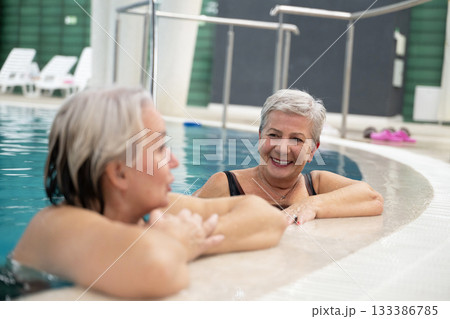 Two smiling senior women enjoying leisure time in an indoor swimming pool, leaning on the poolside. Concept of friendship, wellness, active lifestyle, relaxation, and healthy aging. Two smiling senior women enjoying leisure time in an indoor swimming pool, leaning on the poolside. Concept of friendship, wellness, active lifestyle, relaxation, and healthy aging. 133386785