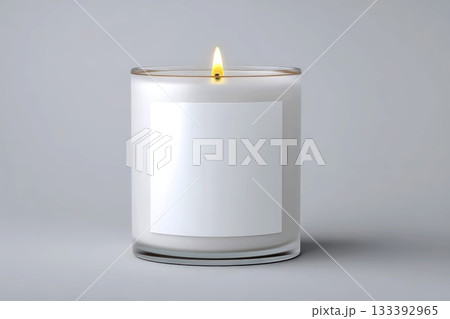 A lit candle in a glass holder with a plain white label, set against a neutral gray background 133392965