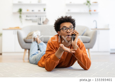 A young man with glasses lies on the floor, talking on his phone with an expressive gesture in a modern living room. 133393401