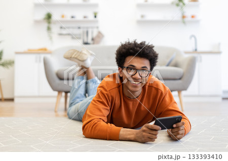 A young man with glasses smiles while lying on the floor and holding a phone in a cozy living room. 133393410