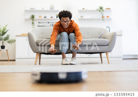 A young man watches a robot vacuum cleaner in his living room, looking excited and amused. 133393415