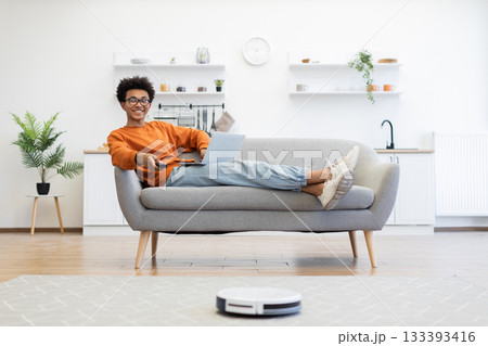A young man relaxes on a couch with a laptop and remote control while a robot vacuum cleans the floor. A young man relaxes on a couch with a laptop and remote control while a robot vacuum cleans the floor. 133393416