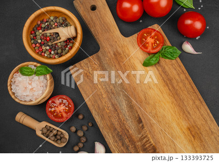 Fresh cooking ingredients tomatoes, basil, spices and garlic arranged around an empty wooden cutting board on dark stone background, top view copy space Fresh cooking ingredients tomatoes, basil, spices and garlic arranged around an empty wooden cutting board on dark stone background, top view copy space 133393725