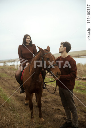 Young romantic couple horse riding, against the background of a field with autumn grass and a lake Young romantic couple horse riding, against the background of a field with autumn grass and a lake 133400587