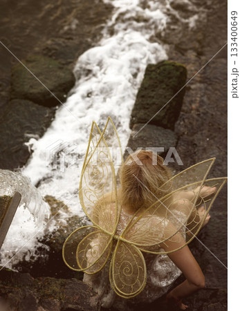 Cute blonde in fairy costume with wings and lace, transparent white dress sits in front of waterfall Cute blonde in fairy costume with wings and lace, transparent white dress sits in front of waterfall 133400649