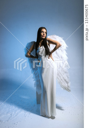 studio photo of blonde in white long dress and angel wings posing on blue background in studio 133400675