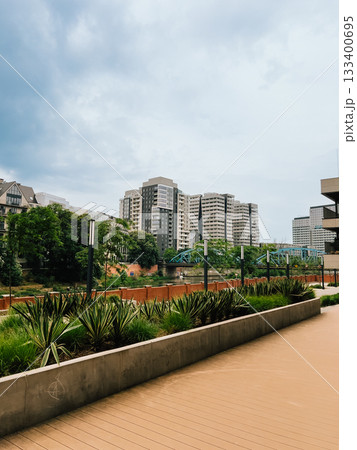 Urban park nestled between high rise residential buildings, featuring wooden walkway, verdant landscape, river passing under arched bridge, embodying contemporary metropolitan tranquility. Living area 133400695