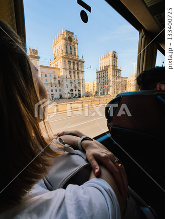 Young woman admiring the stunning Gates of Minsk from window of tourist bus, soaking in vibrant cityscape on bright, sunny day filled with exploration and adventure. Traveler gazing through bus window 133400725