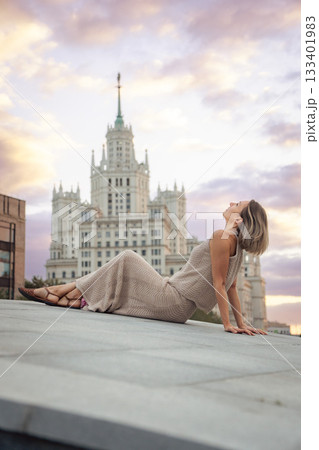 A woman sits on a rooftop at sunset with the Kotelnicheskaya Embankment Building rising behind her near Tessinsky Bridge, surrounded by pastel sky 133401983