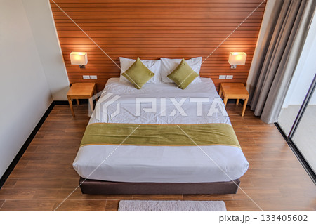 A hotel room with wooden wall paneling and soft lighting displays a large bed with white bedding and green pillows, along with bedside tables holding mounted lamps. A hotel room with wooden wall paneling and soft lighting displays a large bed with white bedding and green pillows, along with bedside tables holding mounted lamps. 133405602