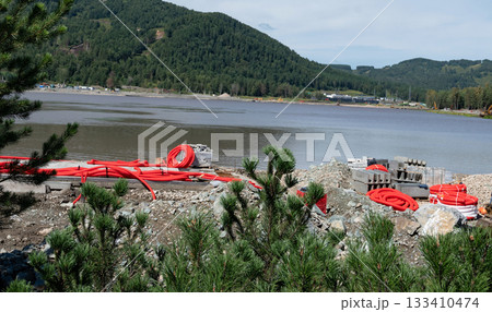 Construction materials by a lake surrounded by green hills. 133410474