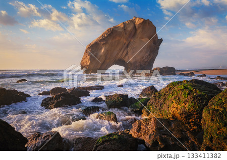 Penedo do Guincho, a large boulder rock arch at Praia da Santa Cruz, Portugal, with ocean waves and sandy beach on sunset Penedo do Guincho, a large boulder rock arch at Praia da Santa Cruz, Portugal, with ocean waves and sandy beach on sunset 133411382
